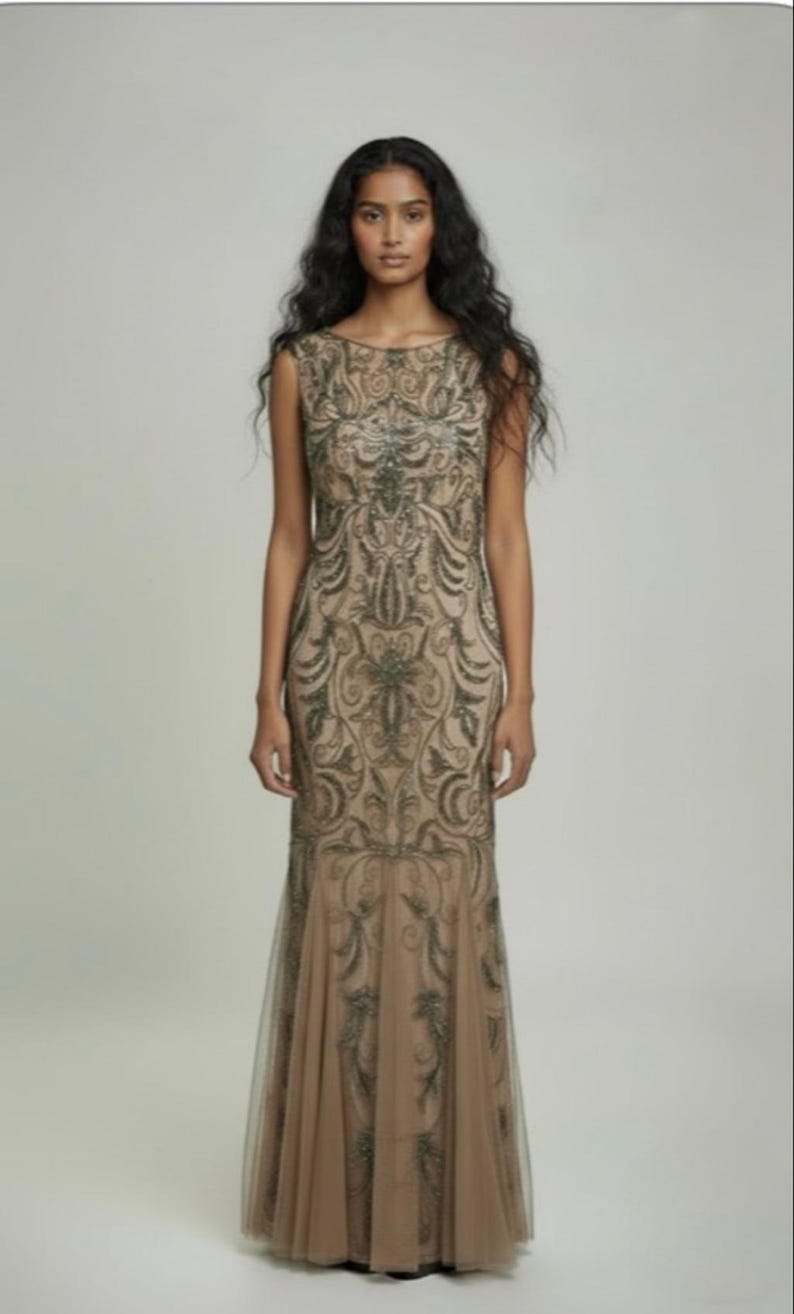 May include: A sleeveless, floor-length gown in a neutral beige hue. The dress features intricate, dark-toned embroidery in a scrolling pattern across the bodice and skirt. The skirt flares slightly at the hem, with a sheer overlay.