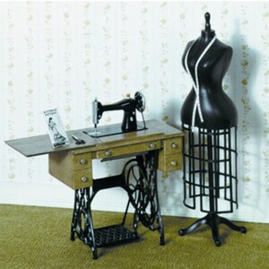 May include: A vintage sewing machine with a black finish sits on a wooden table with drawers. A black dress form with a measuring tape and a sewing pattern are also present. The machine has a decorative cast iron base.