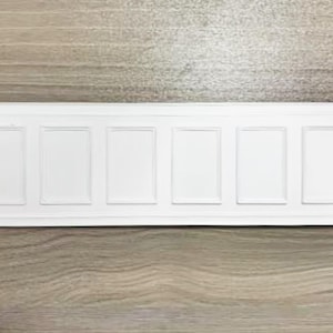 May include: A rectangular white wall panel featuring a wainscoting design with six recessed vertical rectangular sections. The panel has a smooth, matte finish and is framed by horizontal borders along the top and bottom edges of the piece.