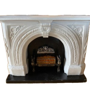 Könnte beinhalten: Ein weißer, verzierter Kaminsims mit schwarzem Innenraum. Der Kaminsims weist detaillierte Schnitzereien auf, darunter Blumen- und Blattmotive. Die Kaminöffnung zeigt einen dekorativen Rost und künstliche Holzscheite.