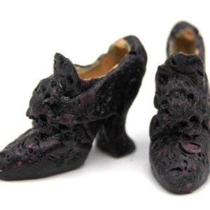 May include: A pair of miniature dark purple shoes with a textured, porous surface and a high heel. The shoes feature a pointed toe and a structured opening, displaying a deep, mottled colour throughout the exterior with a lighter tan interior.