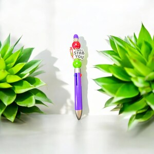 May include: A purple multi-colour pen with a silver tip and a decorative top. The top features a red and green design with the text "I WILL STAB YOU" on a white background. The pen is surrounded by green plants.