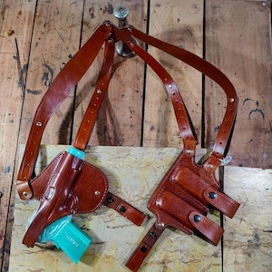 May include: A brown leather shoulder holster with a matching belt and straps. The holster is designed to hold a handgun, with a light blue object inside. The leather is a rich, reddish-brown colour, and the straps are adjustable.