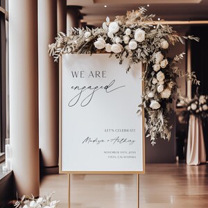 May include: A white sign with a gold frame announcing an engagement. The sign reads "We are engaged" in a script font. Below the text, it says "Let's celebrate" followed by the couple's names, the date, and location. The sign is decorated with white flowers and greenery.