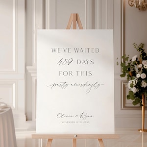 May include: A white wedding sign on a wooden easel. The sign reads "WE'VE WAITED 489 DAYS FOR THIS party accordingly" with the names "Olivia & Ryan" and the date "NOVEMBER 10TH, 20XX" in black script. The sign is in a bright room with floral arrangements.