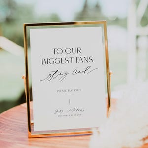 May include: A white sign in a gold frame with the text "To our biggest fans stay cool" and "Please take one!"  The sign also includes the text "Sally and Anthony November 10th 20XX".