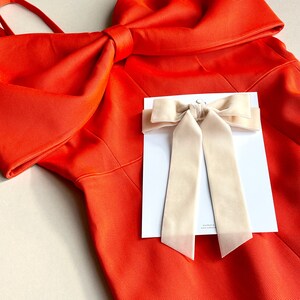 Könnte beinhalten: Ein leuchtend orangefarbenes Kleid mit einer großen Schleife an der Schulter. Ein cremefarbenes Haarschmuck-Accessoire mit Schleife wird auf einer weißen Karte präsentiert und ergänzt die kräftige Farbe des Kleides. Das Kleid hat eine strukturierte Optik.