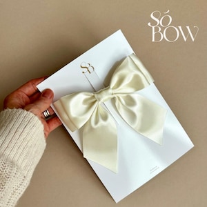 Op de afbeelding: Ivoor satijnen strik op een witte kaart met gouden letters. De kaart heeft het logo "Sö BOW" rechtsboven en een kleiner logo in het midden. De strik is geknoopt en aan de kaart bevestigd.