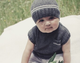 May include: A baby wearing a grey knitted hat with a white stripe and a grey knitted sleeveless top. The baby is sitting on a white blanket and holding a green plant.