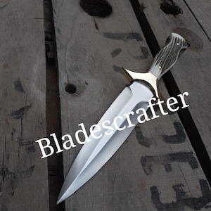 Peut inclure: Un poignard argenté avec une poignée en bois de cerf brun et une garde en laiton. Le poignard est posé sur une surface en bois. Le texte "Bladescrafter" est visible sur la lame.