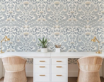 May include: A home office setup featuring a white desk with two matching rattan chairs. The desk has white drawers with gold handles. The background is a floral wallpaper in blue, gray, and beige tones. Two gold desk lamps and a potted plant are on the desk.