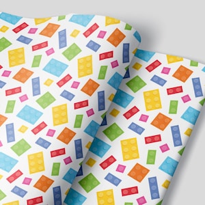 May include: White wrapping paper with a repeating pattern of colorful toy building blocks. The blocks are in various shapes and sizes, including squares, rectangles, and circles.