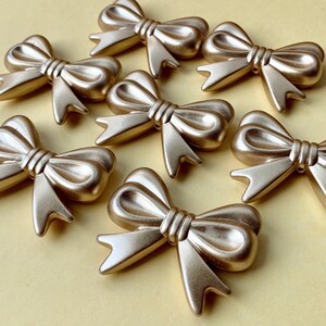 May include: Several gold-colored bow embellishments arranged on a light yellow surface. The bows have a metallic finish and are detailed with realistic folds and a tied center. These decorative bows are suitable for crafts and embellishments.