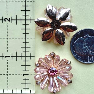 May include: Two flower-shaped embellishments next to a ruler and a US quarter coin. The top piece has metallic gold petals, while the bottom piece features light pink crystal petals with a dark pink center stone and small clear accent crystals.