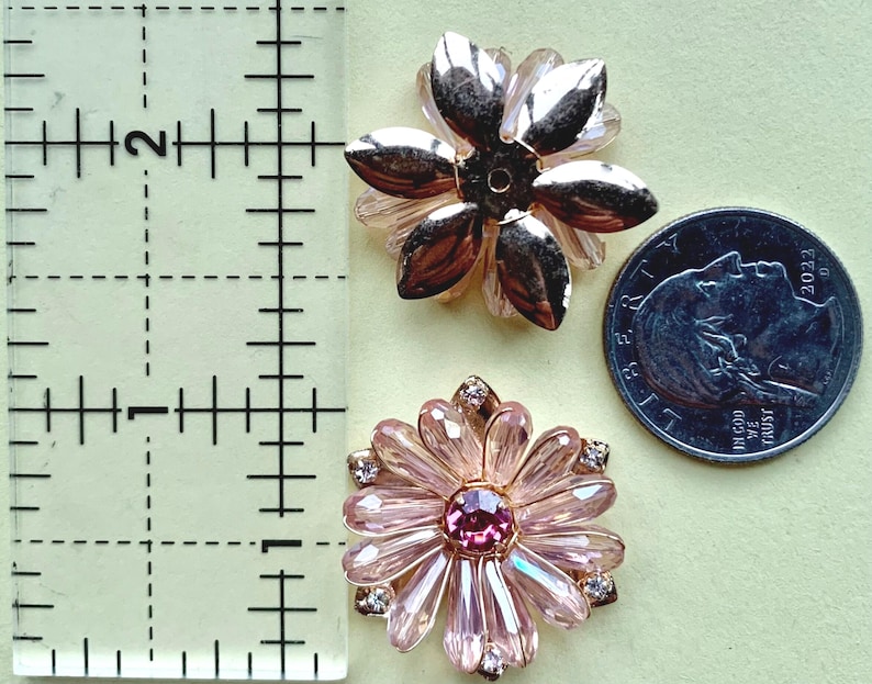 May include: Two flower-shaped embellishments next to a ruler and a US quarter coin. The top piece has metallic gold petals, while the bottom piece features light pink crystal petals with a dark pink center stone and small clear accent crystals.