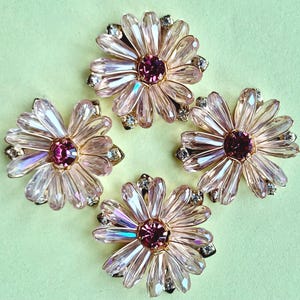 Puede incluir: Cuatro adornos en forma de flor con pétalos de cristal facetado de color rosa claro dispuestos en un patrón circular alrededor de una piedra central de color rosa oscuro. Cada pieza incluye pequeños cristales transparentes en una base dorada.