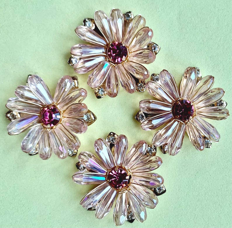 May include: Four flower-shaped embellishments featuring light pink, faceted crystal petals arranged in a circular pattern around a dark pink central stone. Each piece includes small, clear accent crystals set within the gold-toned metal base.