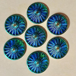 Puede incluir: Siete botones decorativos iridiscentes azules y verdes dispuestos en un patrón circular. Cada botón tiene un elemento central metálico dorado y segmentos radiantes y puntiagudos. Los botones tienen un brillo metálico sobre un fondo neutro.