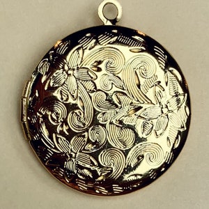 May include: A gold-colored, round locket with a floral and swirl design. The locket has a loop at the top for a chain. The surface of the locket is highly detailed with embossed flowers and swirling patterns.