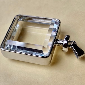 Könnte beinhalten: Ein klarer Kristall-Anhänger mit silbernem Rahmen und silberner Öse. Der Anhänger ist quadratisch und hat ein facettierendes Design.