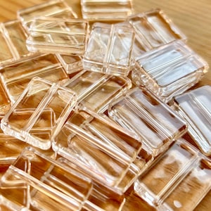 Puede incluir: Un montón de cuentas de vidrio rectangulares transparentes con bordes redondeados. Las cuentas tienen un ligero tono ámbar y están dispuestas sobre una superficie de madera. Las cuentas transparentes son adecuadas para la fabricación de joyas o manualidades.