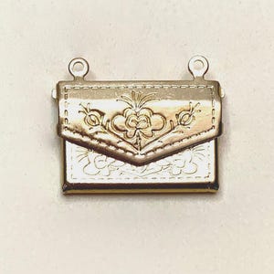 Puede incluir: Un dije de metal dorado con forma de sobre pequeño o bolso con solapa. La superficie presenta un patrón floral grabado y detalles de costura a lo largo de los bordes. Dos pequeños bucles circulares están unidos en la parte superior.