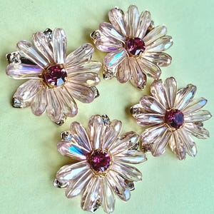 May include: Four flower-shaped brooches featuring light pink, faceted crystal petals arranged in a circular pattern. Each brooch has a deep magenta circular crystal center and small clear accent stones set in a gold-toned metal base.