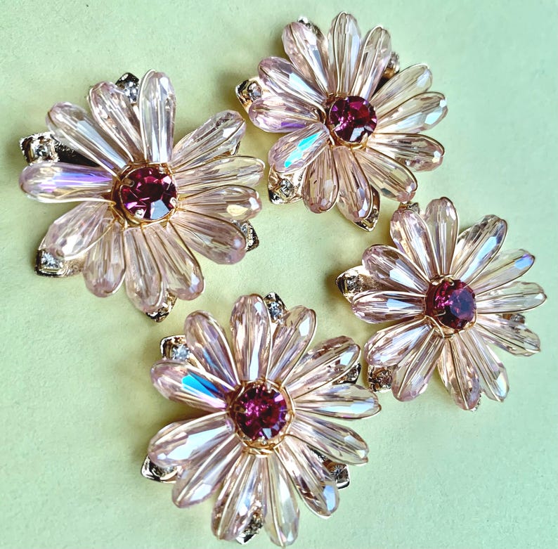May include: Four flower-shaped brooches featuring light pink, faceted crystal petals arranged in a circular pattern. Each brooch has a deep magenta circular crystal center and small clear accent stones set in a gold-toned metal base.