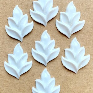 May include: Seven white, leaf-shaped embellishments with a glossy finish. Each leaf has three distinct sections, with a pointed top. The leaves are arranged on a neutral, light brown surface.