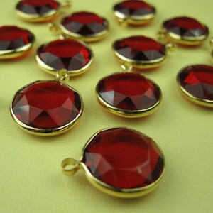 Pode incluir: Um grupo de gemas vermelhas facetadas de vidro com engastes de metal dourado. As gemas estão dispostas em semicírculo sobre uma superfície amarela.