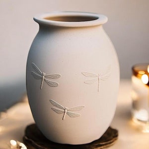 May include: A cream-colored ceramic vase with three embossed dragonfly designs. The vase has a rounded body and a slightly flared rim. A lit candle in a glass holder is in the background, creating a warm, inviting ambiance.