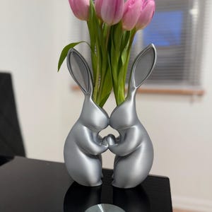 Puede incluir: Un jarr&oacute;n de cer&aacute;mica plateado con forma de dos conejos que se toman de la mano, con tulipanes rosas en el jarr&oacute;n.