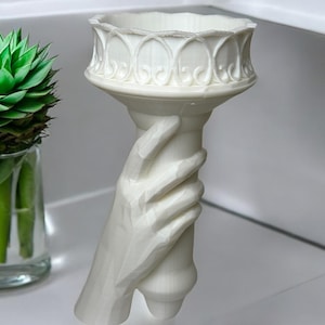 May include: White ceramic sculpture of a hand holding a torch. The torch has a decorative rim and a smooth, cylindrical base.
