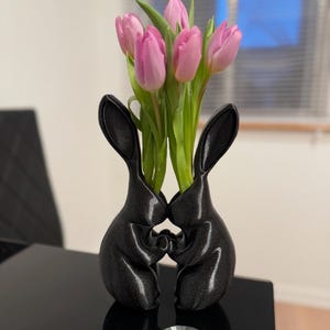 Puede incluir: Un jarr&oacute;n de cer&aacute;mica negro con forma de dos conejos abraz&aacute;ndose, con tulipanes rosas en el jarr&oacute;n.