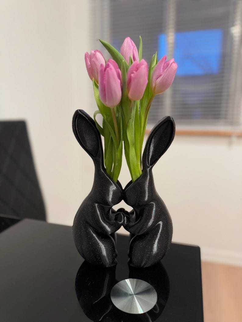Puede incluir: Un jarr&oacute;n de cer&aacute;mica negro con forma de dos conejos abraz&aacute;ndose, con tulipanes rosas en el jarr&oacute;n.