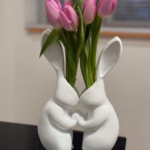 Puede incluir: Un jarr&oacute;n de cer&aacute;mica blanca con forma de dos conejos que se toman de la mano, con tulipanes rosas en el jarr&oacute;n.