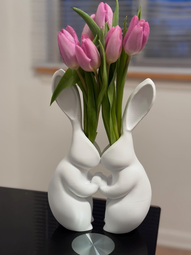 Puede incluir: Un jarr&oacute;n de cer&aacute;mica blanca con forma de dos conejos que se toman de la mano, con tulipanes rosas en el jarr&oacute;n.
