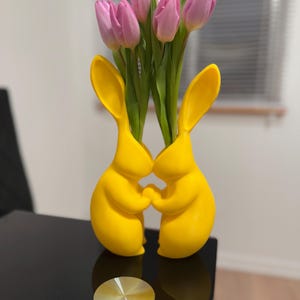 Puede incluir: Dos figuritas de conejo de cer&aacute;mica amarilla que se toman de la mano, formando un jarr&oacute;n. El jarr&oacute;n est&aacute; lleno de tulipanes rosas.