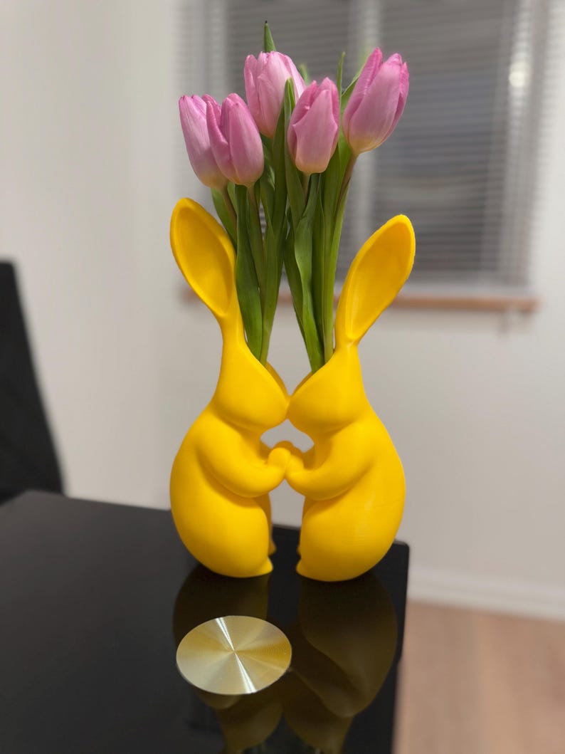 Puede incluir: Dos figuritas de conejo de cer&aacute;mica amarilla que se toman de la mano, formando un jarr&oacute;n. El jarr&oacute;n est&aacute; lleno de tulipanes rosas.