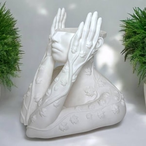 May include: White ceramic planter shaped like a human bust with hands covering the face. The hands and bust are decorated with raised floral designs.