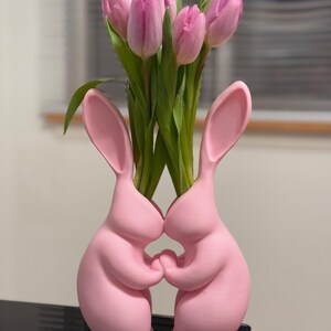 Puede incluir: Jarr&oacute;n de cer&aacute;mica rosa en forma de conejo con dos conejos que se toman de la mano, sosteniendo un ramo de tulipanes rosas.
