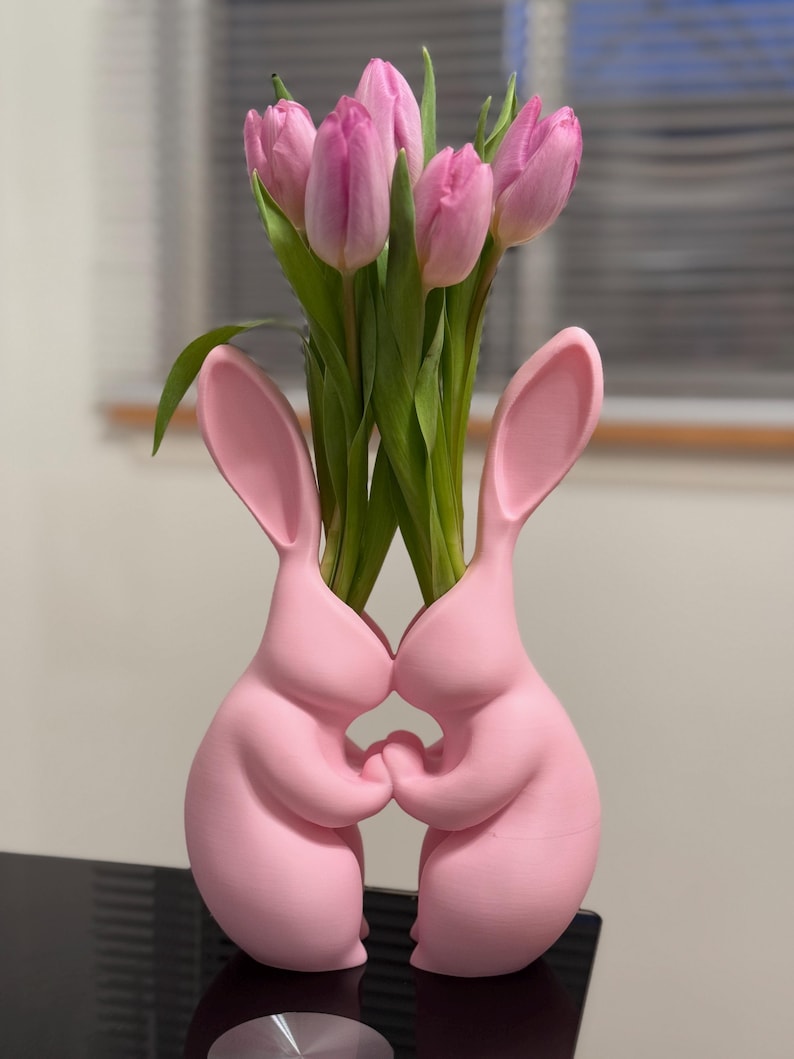 Puede incluir: Jarr&oacute;n de cer&aacute;mica rosa en forma de conejo con dos conejos que se toman de la mano, sosteniendo un ramo de tulipanes rosas.