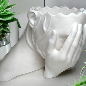 Peut inclure: Pot en céramique blanche en forme de personne avec les mains sur le visage. Le pot est conçu pour contenir une petite plante ou une succulente.