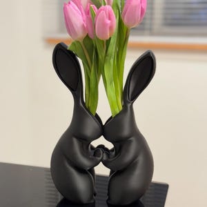 Puede incluir: Jarr&oacute;n de cer&aacute;mica negro con forma de dos conejos que se toman de la mano, con tulipanes rosas en el jarr&oacute;n.