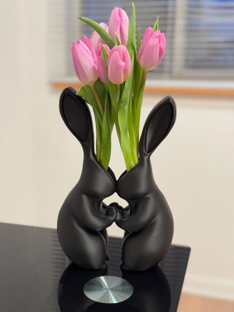 Puede incluir: Jarr&oacute;n de cer&aacute;mica negro con forma de dos conejos que se toman de la mano, con tulipanes rosas en el jarr&oacute;n.