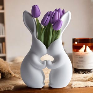 Puede incluir: Jarrón de cerámica blanco con forma de dos conejos que se toman de la mano, con un ramo de tulipanes morados en su interior.