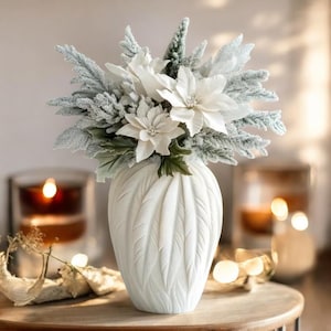May include: White ceramic vase with a textured, leaf-like design, filled with a bouquet of white poinsettia flowers and frosted greenery.