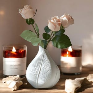 May include: A white, leaf-shaped vase holds pink roses with green leaves. Two lit candles in glass jars flank the vase. The candles have a dark red wax and white labels. The scene is set on a wooden surface.