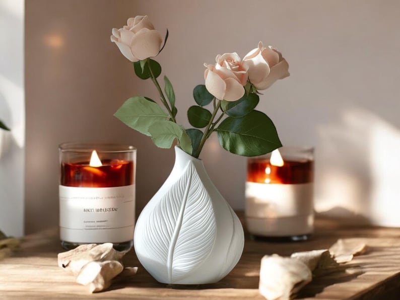 May include: A white, leaf-shaped vase holds pink roses with green leaves. Two lit candles in glass jars flank the vase. The candles have a dark red wax and white labels. The scene is set on a wooden surface.