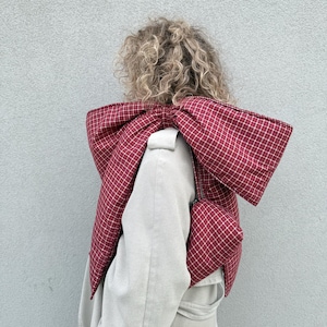 Könnte beinhalten: Ein rot-weiß karierter, schleifenförmiger Schal, getragen über einem hellen Mantel. Der Schal ist aus gestepptem Stoff gefertigt und hat eine passende kleine Tasche. Ein einzigartiges Modeaccessoire.